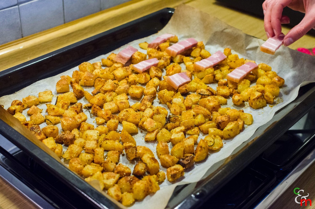 Dupa 15-20 de minute, scoateti tava, amestecati cartofii cu o paleta si adaugati feliile de bacon peste cartofi. Bagati tava la cuptor pentru inca 10-15 minute.